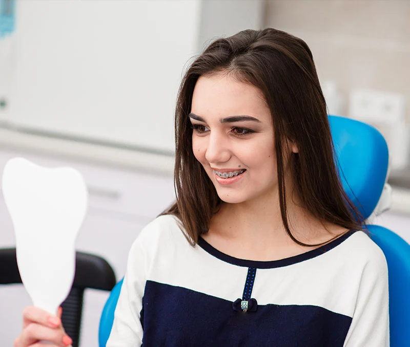 Smiling teenage patient wearing braces holding a hand mirror